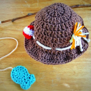 May include: A brown crocheted hat with a white brim and colorful yarn accents. The hat is sitting on a wooden surface with a blue crocheted fish and a stick with string attached.