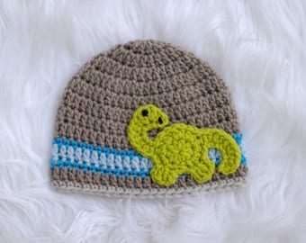 May include: A crocheted baby hat in shades of brown, with a light blue and blue stripe near the brim. A lime green dinosaur applique is on the front. The hat is made of soft yarn and is suitable for a baby.