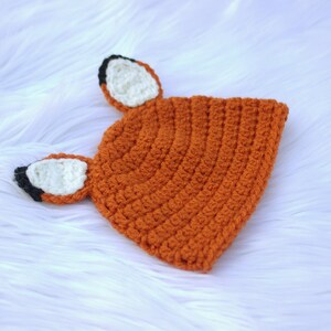 May include: A crocheted fox hat with orange, white, and black accents. The hat has two fox ears with white tips and black outlines.