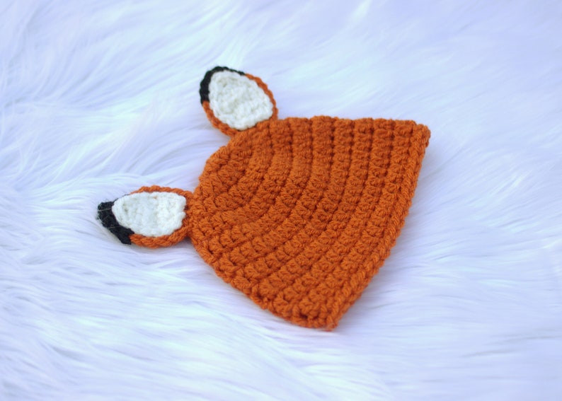 May include: A crocheted fox hat with orange, white, and black accents. The hat has two fox ears with white tips and black outlines.