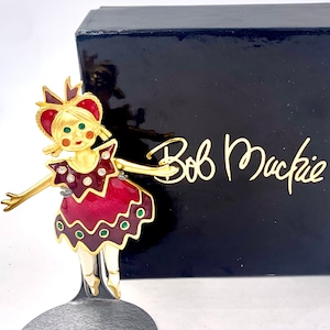 Puede incluir: Una figurita dorada de una bailarina que lleva un vestido rojo y verde con un sombrero blanco y verde. La figurita está sobre un soporte negro y la caja en la que venía dice "Bob Mackie".