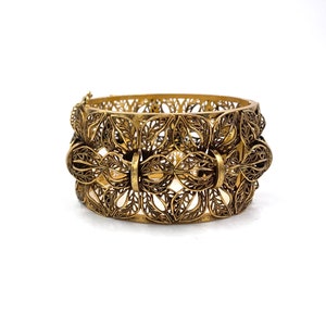 May include: A gold-toned filigree bracelet with a delicate leaf pattern. The bracelet is made of thin metal and has a hinged clasp.
