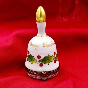 Könnte beinhalten: Eine weiße Porzellan-Weihnachtsglocke mit goldenen Akzenten. Die Glocke trägt den goldenen Schriftzug "Merry Christmas" sowie Illustrationen von Stechpalmen und roten Beeren. Sie hat einen goldenen Klöppel und eine goldfarbene Spitze.