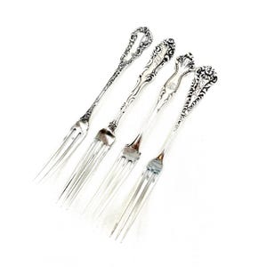 May include: Four silver-toned metal forks arranged diagonally. Each fork features a distinct ornate handle with intricate scrollwork and floral detailing. The tines are long and slender, and some handles display engraved monogram initials in cursive script.