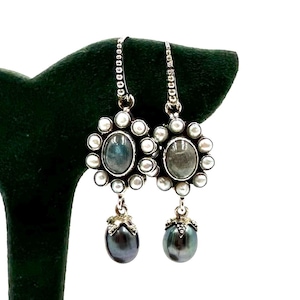 May include: A pair of silver earrings with oval, iridescent stones surrounded by small pearls. Each earring has a teardrop-shaped, dark gray pearl pendant. The earrings are displayed on a dark green velvet surface.
