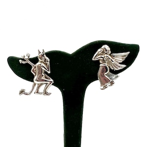 May include: A pair of silver-toned earrings. One earring depicts a devil with a trident, the other an angel with wings. The earrings are displayed on a black velvet jewelry stand against a white background.