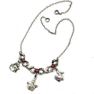 May include: Silver-toned necklace featuring bow-shaped links with red accents and dangling charms. Charms include a rabbit, a duck, and a seated animal. The necklace has a delicate chain and a clasp closure.