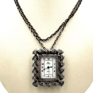May include: Silver-toned watch necklace with a rectangular watch face. The watch face has black numerals and hands against a white background. The watch is framed by a decorative floral border and hangs from a double chain.