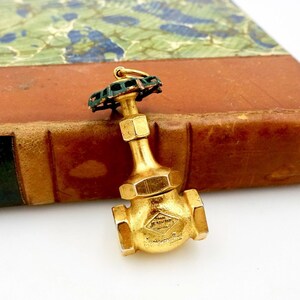 May include: Gold-toned metal charm shaped like a water valve with a green and copper colored top. The valve has the text "Jenkins Bros" on the side.