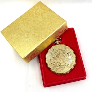 May include: A gold-colored pocket watch with an ornate floral design, resting in a red velvet-lined box. The watch is round with a scalloped edge and a detailed engraved scene. The watch is presented in a gold box with a floral pattern.