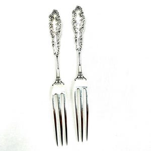 May include: Two silver-colored metal forks featuring ornate, embossed scrollwork patterns on the handles. Each fork has four long, slender tines and a decorative neck section. The metal surface displays a polished, reflective finish throughout.