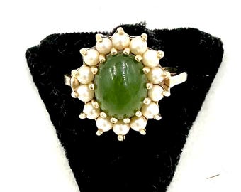 Könnte beinhalten: Ein goldener Ring mit einem ovalen, grünen Edelstein in der Mitte, umgeben von kleinen weißen Perlen. Der Ring liegt auf einem schwarzen Samtuntergrund. Das Design ist klassisch und elegant, im Stil von Vintage-Schmuck.