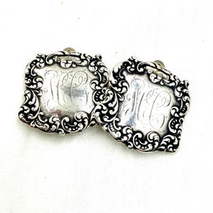 May include: A pair of antique silver-toned cufflinks. Each cufflink features a rectangular shape with ornate scrollwork around the edges. The center of each cufflink has an engraved monogram. The cufflinks are displayed on a white surface.