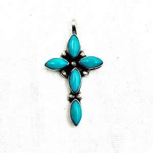 Pode incluir: Um pingente em forma de cruz feito de metal prateado, com quatro pedras de cor turquesa lapidadas em marquise dispostas em cruz. O centro da cruz é acentuado com uma pequena conta prateada e possui uma argola de metal no topo para pendurar.