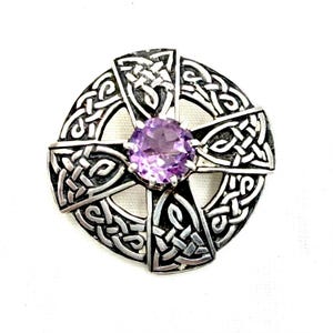 May include: A circular silver brooch featuring an intricate interlaced knotwork pattern forming a cross shape. At the center of the cross is a round, faceted, light purple amethyst gemstone held in place by four small metal prongs.