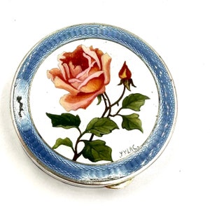 May include: A silver compact with a blue enamel border and a hand-painted rose design. The compact is signed "YVLAC."