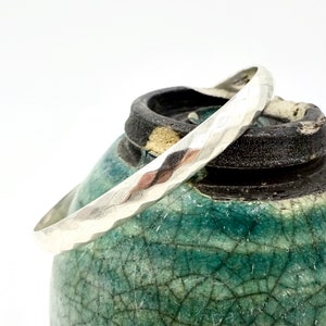 May include: A hammered silver bangle bracelet. The bracelet has a textured surface and is displayed on a teal and black ceramic pot. The bangle is a simple, elegant piece of jewelry.