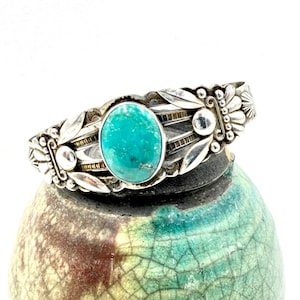 May include: Silver cuff bracelet featuring an oval turquoise stone. The bracelet has a detailed design with leaf and floral accents. The turquoise stone is a vibrant teal color, set in a silver band. The bracelet is displayed on a ceramic object.