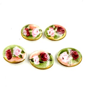 Puede incluir: Cinco botones ovalados de cerámica pintados a mano. Cada botón presenta un diseño de rosas rosas y rojas sobre un fondo verde y melocotón, con un borde dorado. Los botones están dispuestos sobre una superficie blanca.