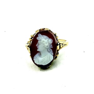 Könnte beinhalten: Ein goldener Ring mit einem ovalen Cameo, der das Profil einer weißen Frau auf einem dunkelroten Hintergrund zeigt. Der Ring hat eine verzierte Goldfassung und ein Band mit dekorativen Details an den Seiten.