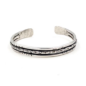 May include: A silver bracelet with a triple band design. Each band has a textured pattern of small circles and triangles.