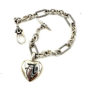 Puede incluir: Un brazalete de plata con un dije en forma de corazón. El dije tiene una letra 'J' estilizada grabada en él. El brazalete tiene un cierre de palanca.