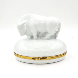Könnte beinhalten: Weiße Porzellan-Schmuckdose mit einer detaillierten Bullenskulptur auf dem Deckel. Die Dose hat einen goldenen Scharnierdeckel.