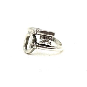 Pode incluir: Um anel prateado com um design em forma de chave. O anel apresenta uma chave com as letras "AOO" na cabeça da chave. O anel é feito de metal brilhante e está sobre um fundo branco.