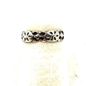 May include: A silver-colored ring with a repeating floral design. The ring has a textured surface and a band of connected flower-like shapes. The ring is displayed on a white, textured surface.