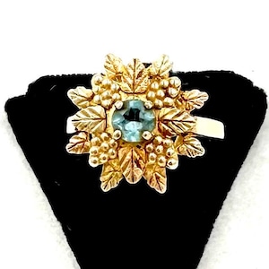 May include: A gold-toned ring featuring a central, round, light blue gemstone. The ring's design includes a cluster of gold-colored leaves and grape-like details surrounding the gemstone, creating a floral or botanical theme. The ring is set against a black background.