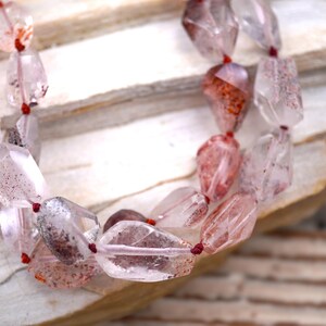 May include: A necklace featuring faceted, translucent quartz beads with internal red and brown mineral inclusions. The irregular, polished stones are strung together on a thin, dark red cord, creating a multi-layered appearance with varied shapes.