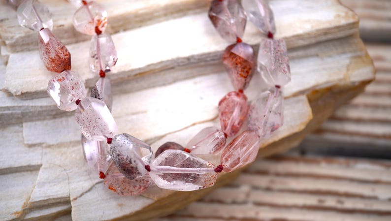 May include: A necklace featuring faceted, translucent quartz beads with internal red and brown mineral inclusions. The irregular, polished stones are strung together on a thin, dark red cord, creating a multi-layered appearance with varied shapes.