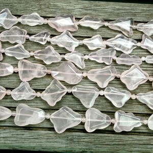 May include: A strand of pink rose quartz beads, each bead is faceted and shaped like a teardrop. The beads are strung on a clear elastic cord and are arranged in a staggered pattern.
