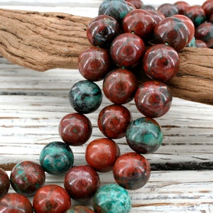 Puede incluir: Hileras de cuentas grandes y redondas en tonos rojos y verde azulado. Las cuentas tienen un aspecto veteado y están ensartadas. Un trozo de madera flotante descansa en el fondo, con una superficie de madera blanca.