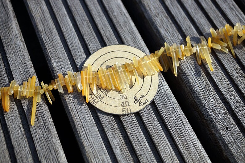 May include: A string of yellow and clear crystal beads laid out on a wooden surface. The beads are arranged in a line and are all different shapes and sizes. A round target with the words "Sparkle Little" and numbers 30, 40, and 50 is visible in the background.