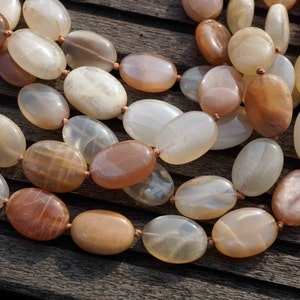 May include: A necklace featuring large, oval-shaped beads in shades of cream, peach, and brown. The beads are strung together with a thin, light-brown cord. The necklace is displayed on a dark wooden surface, showcasing the natural tones of the gemstones.