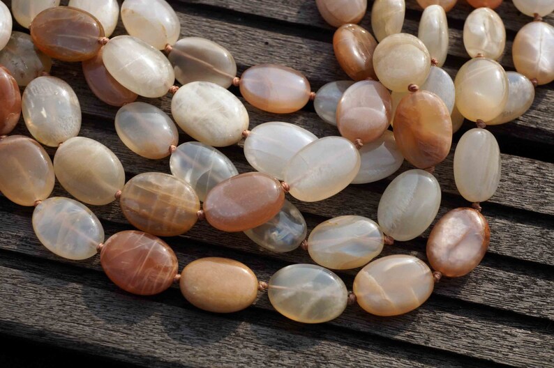May include: A necklace featuring large, oval-shaped beads in shades of cream, peach, and brown. The beads are strung together with a thin, light-brown cord. The necklace is displayed on a dark wooden surface, showcasing the natural tones of the gemstones.