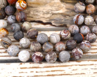 May include: Close-up of a necklace featuring a variety of round, patterned beads in shades of brown, gray, and cream. The beads have unique veining and textures, strung together on a gold-colored cord. The necklace is displayed on a wooden surface.