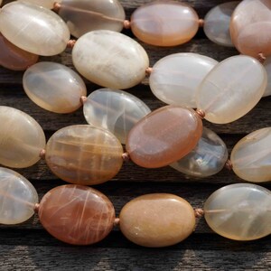 May include: A close-up of a necklace featuring large, oval-shaped beads in shades of cream, peach, and brown. The beads are strung together with a thin, light brown cord. The necklace is displayed on a wooden surface.