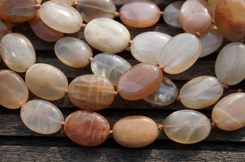 May include: A close-up of a necklace featuring large, oval-shaped beads in shades of cream, peach, and brown. The beads are strung together with a thin, light brown cord. The necklace is displayed on a wooden surface.