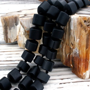 Puede incluir: Una hebra de cuentas cuadradas de acabado mate negro. Las cuentas están enhebradas en un cordón negro y están dispuestas en un patrón escalonado.