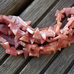 May include: A strand of pink triangle-shaped gemstone beads. The beads are made of a pink and white striped material.