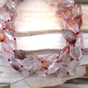 May include: A multi-strand necklace composed of faceted, clear quartz beads featuring internal reddish-brown inclusions. The beads are strung on a thin, dark red cord, creating a layered appearance with a translucent, crystalline texture throughout the piece.