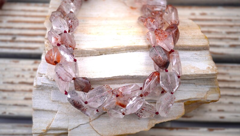 May include: A multi-strand necklace composed of faceted, clear quartz beads featuring internal reddish-brown inclusions. The beads are strung on a thin, dark red cord, creating a layered appearance with a translucent, crystalline texture throughout the piece.
