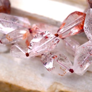 May include: A strand of faceted, translucent quartz beads featuring internal red and brown mineral inclusions. The irregular, polished stones are strung together with a thin, dark red cord, creating a multi-faceted, light-reflecting appearance.