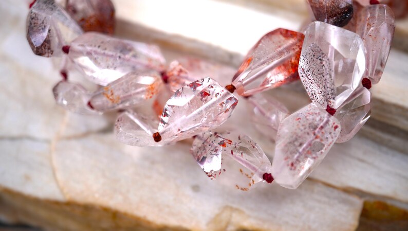 May include: A strand of faceted, translucent quartz beads featuring internal red and brown mineral inclusions. The irregular, polished stones are strung together with a thin, dark red cord, creating a multi-faceted, light-reflecting appearance.