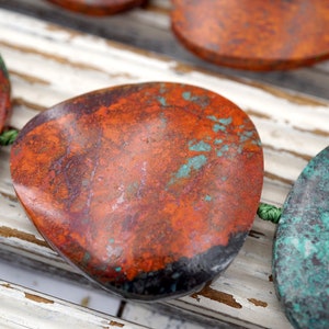 Puede incluir: Primer plano de varias piedras grandes, de forma irregular y aspecto moteado. Las piedras tienen un color base marrón rojizo con parches de color turquesa y negro. Están ensartadas con un cordón verde, lo que sugiere que son cuentas o parte de una joya más grande.