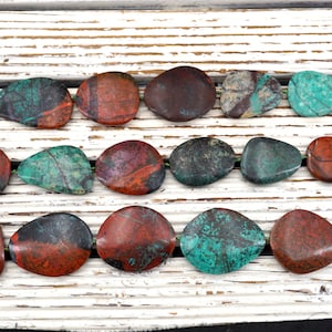 Puede incluir: Una hebra de piedras lisas y pulidas en forma de lágrima con una mezcla de colores rojo, marrón, verde y negro. Las piedras están dispuestas en una fila sobre una superficie de madera blanca.