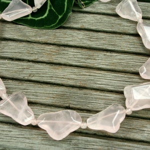 May include: A necklace made of pink rose quartz beads, each bead is a unique, irregular shape. The necklace is laid out on a wooden surface.