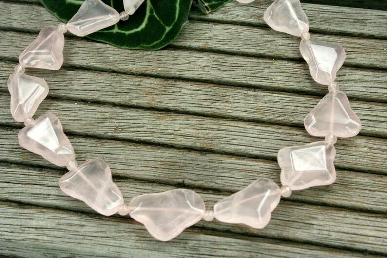 May include: A necklace made of pink rose quartz beads, each bead is a unique, irregular shape. The necklace is laid out on a wooden surface.